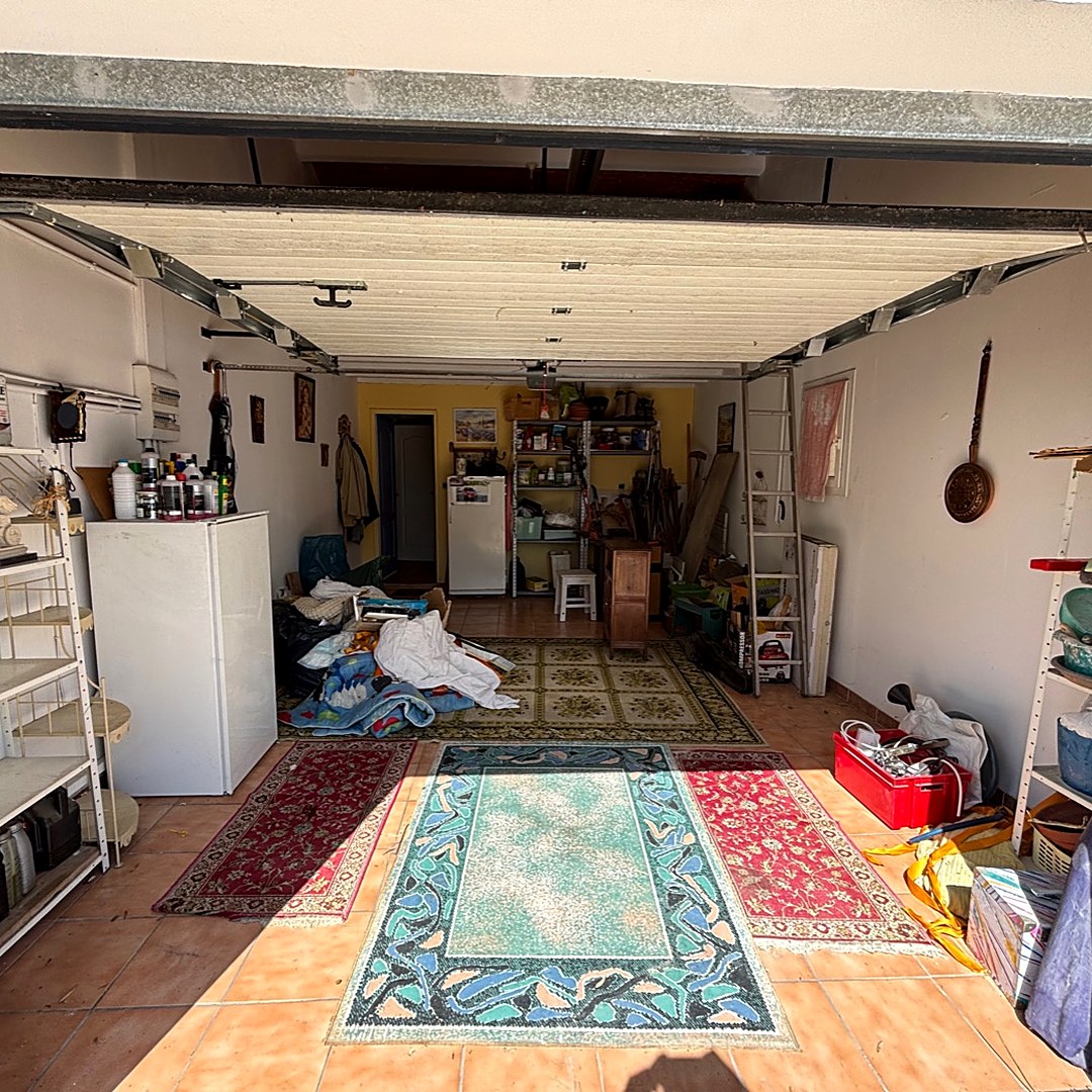 Le même garage avant intervention, encombré de tapis, meubles, étagères et cartons.