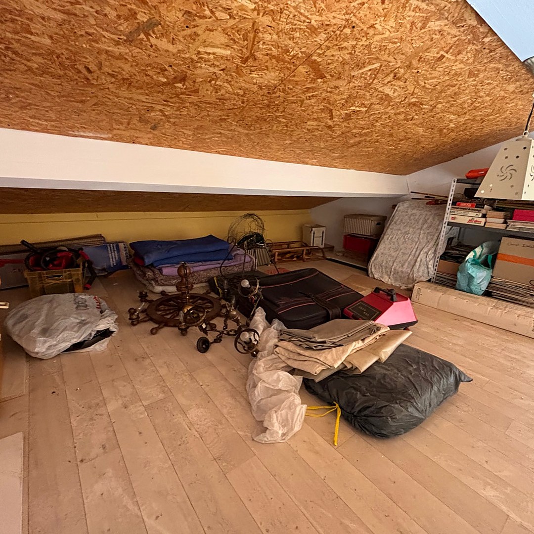Le même grenier avant intervention, encombré de linge, valises, luminaires anciens et cartons.